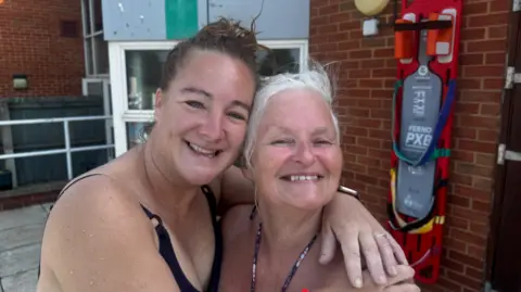 Two women in swimming costumes, hugging, one with short brown hair and the other with short grey hair. They are standing outdoors in front of a brick wall and a large glass door