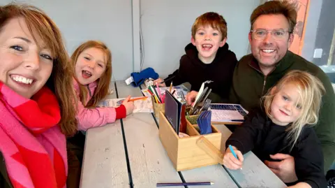 Katie and Tom's family are sitting around a table in a restaurant. Mother Katie and father Tom are with their three children, a boy and two girls. 