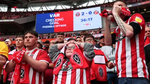 Dozens of Sheffield United fans, mostly dressed in red and white club shirts, look on in anguish in the lower tier behind the goal at Wembley Stadium during the Championship play-off final moments before their second goal against Sunderland was disallowed by VAR for offside.