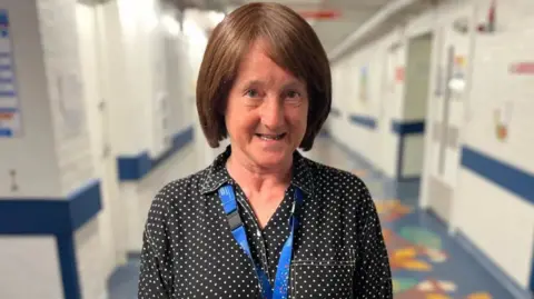 Somerset NHS Foundation Trust Helen Parfitt wears a black polka dot shirt and blue lanyard in a hospital corridor. 