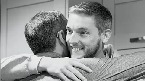 Black and white image of two men hugging. One of the men (right) who is facing the camera has a grey beard and hair and is wearing a grey hoodie with the logo Derian Dads on it. He is smiling. 