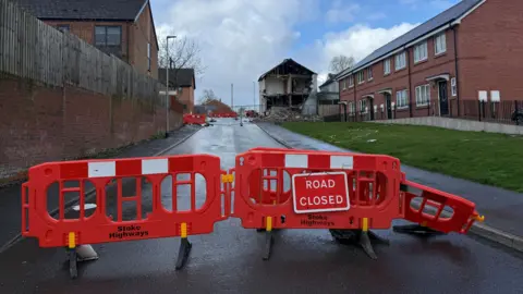 Orange safety barriers block a road where further along there is a partially-demolished building. There are houses and pavement either side of the road and a grass verge on the right hand side.