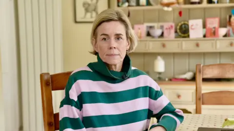 Ant Saddington/BBC Kate Speirs, sat in a room, looking straight at the camera, wearing a pink and green top, she has short blonde hair and earrings in both ears. She has her hand on a table, and is sat on a pine chair. A dresser is behind her with cards and ornaments on it. 