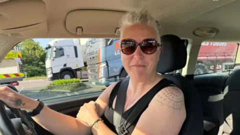 Lili Sheppard/BBC A woman wearing sunglasses and a sleeveless black top is sitting in the driver's seat of a car. Lorries are in a car park in the background.