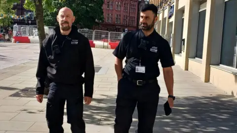 Sheffield Council Two men wearing black uniforms are walking through Sheffield city centre. One has a bald head and brown goatee beard, the other has black hair and a beard
