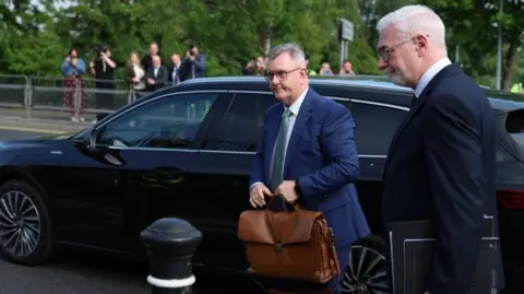 PA Media Jeffrey Donaldson, with short grey hair, wearing a navy suit, white shirt, green tie and glasses. He is holding a brown briefcase and is standing beside a black car. His solicitor John McBurney is standing to the right of him. He has short white hair and stubble, and is wearing a white shirt and black suit. 