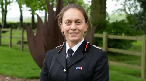 A woman wearing ceremonial fire service uniform is standing outdoors in a garden with trees and fence behind her. She has brown hair and is smiling for the photograph.