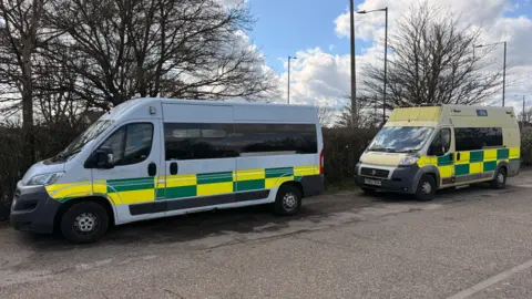 Supplied Two large ambulances parked behind one another. One is white with a yellow and green pattern, while the other is yellow with a yellow and green pattern. 