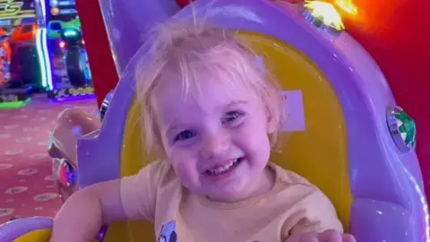 PA Media Smiling female toddler with blonde hair in an amusement arcade. She appears to be seated on a ride. She is wearing a t-shirt depicting Minnie Mouse.