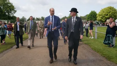Sam Giddens/PA Media HRH Prince of Wales, Prince William, being escorted around the Royal Norfolk Show. The prince is wearing blue trousers and a blue jacket, and he is gesticulating with his hands. His guide is wearing a suit and bowler hat. Other people are in the vicinity.