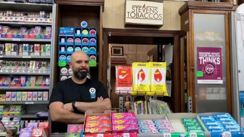 Hsin-Yi Lo/BBC A man in a black t-shirt is standing behind a counter full of lollies. There are tobacco products on a shelf behind him.