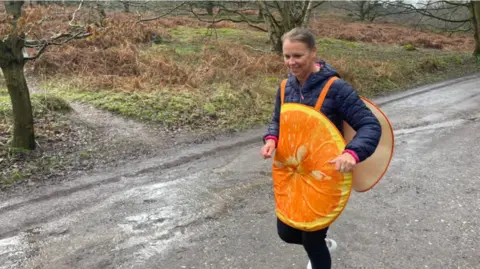 A Sally Orange dressed in an orange fruit costume with a dark blue jacket underneath.