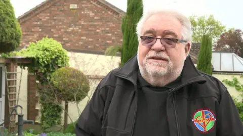 A man with white hair, a grey beard and glasses speaks in a garden. He wears a black jacket with the Lincolnshire Emergency Blood Bikes Service logo with a Lincolnshire flag and heart. 