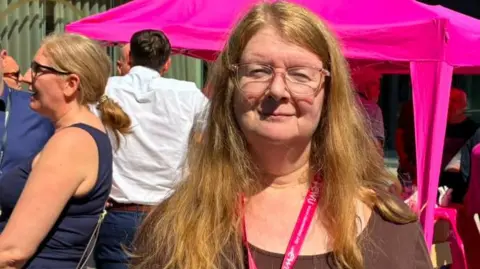 Lesley Wood with long blond hair and glasses wearing a brown t-shirt. She stands in front of a handful of protesters in Liverpool city centre. She is smiling.
