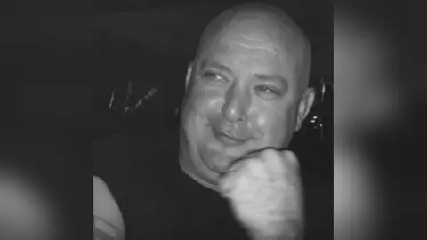 A bald man with a black T-shirt smiles cheekily in a black and white photo. 