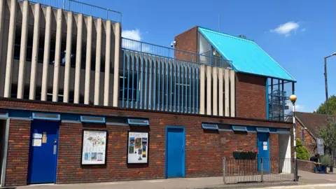 Drovers Way toilet block, showing a large brick building, with blue doors, blue windows and cars parked above. There is a street lamp to the right and a street crossing.
