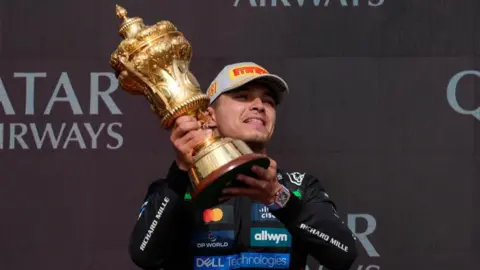 PA Media Lando Norris in a black racing suit and cap holding a gold trophy aloft