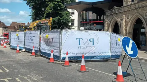 Fiona Callow/BBC A view of a road, cordoned off with fluorescent orange cones and metal fencing. In the foreground, a blue circular sign can be seen with an arrow directing traffic around the area. In the background a digger can be seen, and a stone building in the background with archways.