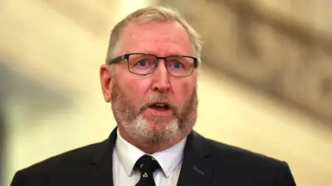 Getty Images Doug has short grey hair with a ginger and grey beard and moustache. He is wearing a black blazer, white shirt and tie.