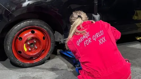 Hannah Simpson A woman in red overalls is using a jack to lift a black car. It has black tyres and red alloys.