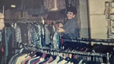 Christine's Wool Shop A woman with dark short hair is wearing a grey coat and is holding hangers with dresses. She is standing next to clothes racks with dresses and suits. 
