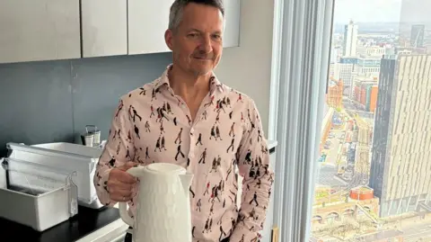 Ken Price in a patterned shirt holds a kettle in his kitchen. HIs window shows Deansgate.