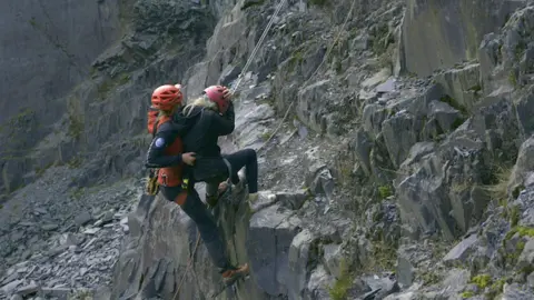 Darlun/BBC Cymru Wales depicts the image two people on a very slope cliff. Both are women with helmets, contestants, and ropes for safety. The savior wears a dark orange jacket, an orange helmet, and long -distance walking shoes. They support the second person, who wears white coaches, a red helmet and has her head. The environment is characterized by rough rock formations and sharp tendency, as the two women are carefully from the rocky face.