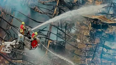 Two firefighters on an aerial platform spraying water over the what is left of the roof of a building. Only the blackened frame of the roof remains.