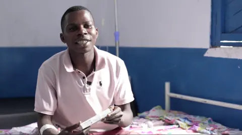 Sadiki Bichichi Aristide sits on a hospital bed, carrying a pharmaceutical package