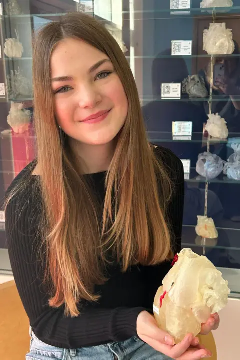 Rebecca Bannister Jasmine in a black top and blue jeans holding a white rubber heart model and smiling at the camera.