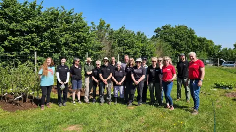 East Devon District Council A large group of people are stood in a field on a sunny day with small trees planted behind them.