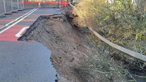 The landslip on Haslingden Old Road showing a chunk of the road collapsed so soil and foliage  can be seen