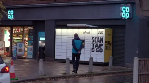 A teacher stands outside a Co-op supermarket beside an InPost parcel locker on a wet street, with a white car passing in the foreground.