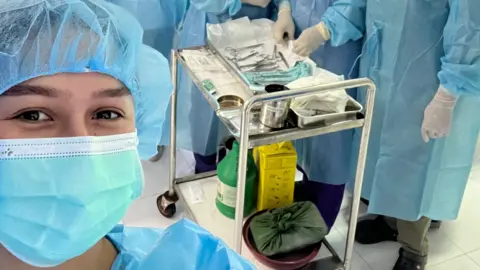 Angela Aumonier lleva una mascarilla azul, una redecilla para el pelo y un mono de papel azul. Detrás de ella hay un carrito plateado con material médico y un grupo de personas vestidas igual.