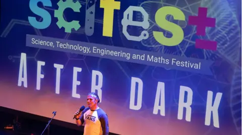 A man in a white shirt with dark sleeved stands in front of a screen holding a microphone. The screen says 'Scifest After Dark' and 'Science, Technology, Engineering and Maths Festival'