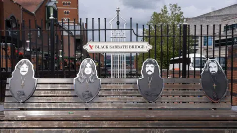 Getty Image Birmingham, Black Sabbath Bridge in United Kingdom. It has pictures of Tony Iomi, Bill Ward, Geyser Butler and Ozi Osbourne. There is a gate behind it and there is a sign that reads Black Sabbath Bridge. 