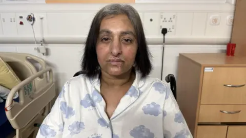 Manjit in a hospital room dressed in patterned pyjamas, with the back of a wheelchair partially visible. Surrounding furniture includes a hospital bed and a wooden cabinet. She is in hospital. 