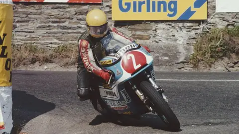 Getty Images Joey Dunlop racing a white and blue bike with the number two in a red circle on the front. He is wearing a black racing suit with red and white detailing on the sleeves. He is wearing a yellow helmet. Behind him is the stone wall with advertisement boards on it.