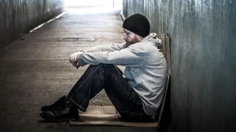 Getty Images A man wearing a black hat, grey top and dark jeans and shoes, sits on a piece of cardboard. He is in a subway tunnel, sitting with his back up against the tunnel's wall