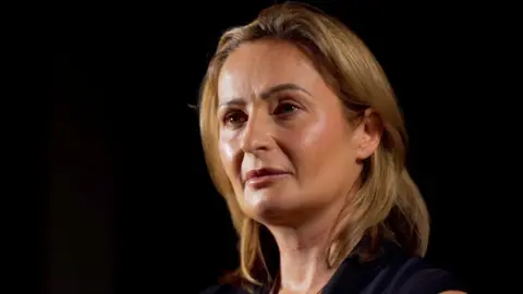 BBC Rhiannon Creak looks off-camera at an interviewer. She has shoulder length blonde hair. She is wearing a black sleeveless dress and necklace. She is set against a plain black background.