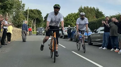 Two men wearing white t-shirts on bikes as they get clapped on at the finishing line on a road in Ballaugh.