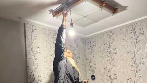 Yulia, wearing her quilted coat, stands on a sofa as she hangs a battery-powered bulb from the ceiling in her home