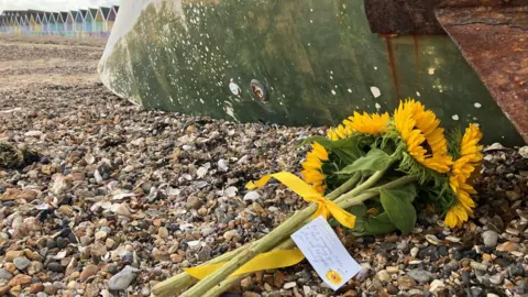 Lewis Adams/BBC A bouquet of yellow sunflowers with a handwritten note attached laid on the pebbly beach. A row of colourful beach huts are pictured in the background.