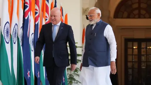 ANI Prime Minister of New Zealand Christopher Luxon (in black and white suit) and Prime Minister of India Narendra Modi (in blue and white Indian attire), with flags of the two countries in the background.
