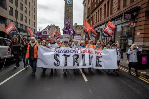 A group of workers protesting as they walk down a street, holding several banners and signs - including one saying GLASGOW EQUAL PAY STRIKE.