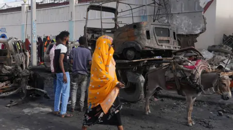 BBC/Mohamud Abdisamad A woman walks past the scene of the attack, with burnt-out cars behind her