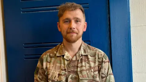 A man with short brown curly hair sweept to the side of his forehead with a ginger short beard and moustache is smiling into the camera and standing against a blue door. He is wearing a camo green and brown uniform.
