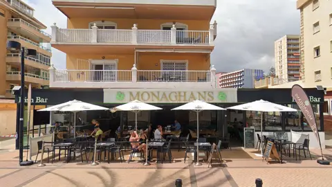Google A bar with outdoor seating and umbrellas below an apartment block with the sing saying Monaghans with a shamrock design.