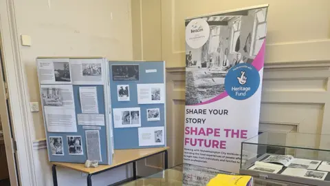 Wolverhampton Archives An exhibition with a tall board that reads "share your story shape the future". There are boards and glass cabinets next to it with documents and black and white photographs included