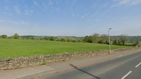 Google A large grass field bordered by a stone wall and roadside pavement, with rolling countryside visible in the distance.